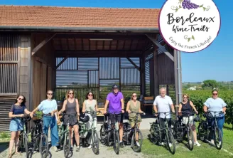 A group of ten cyclists with their bikes standing in front of a modern wooden building overlooking vineyard landscapes in Bordeaux, France.