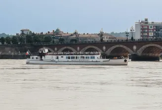 A white river cruise boat named 'Yacht de Bordeaux' sails on the Garonne River in front of the historic Stone Bridge, with 18th-century buildings and architecture visible along the waterfront of Bordeaux, France.