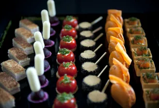 Fine pastry and gourmet appetizers display featuring layered cakes, white cylindrical desserts, red tomato tartlets, black canapés with cream, glazed orange pastries, and herb-topped squares on a black serving board