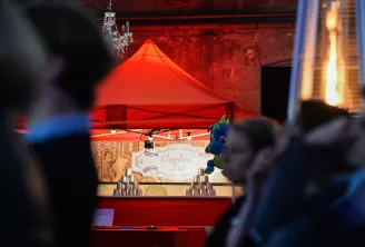 A festive market stall with a red tent canopy displaying 'Chamboul Tout' sign, decorated with a colorful peacock figure, surrounded by stacked cups and visitors in Bordeaux's Christmas market