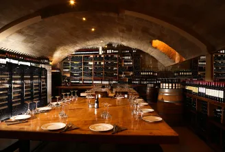 Underground wine cellar with arched stone ceiling, long wooden dining table set for service, wine glasses and place settings, extensive wine bottle storage shelves lining the walls, warm ambient lighting.