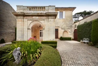 Stone entrance of the Couvent des Jacobins in Bordeaux featuring an arched doorway with decorative molding, manicured hedges, flowering plants, and a brick courtyard surrounded by historic stone buildings