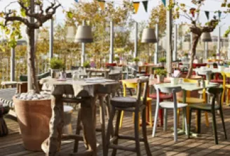 Outdoor terrace of Mama Shelter Bordeaux with wooden tables and chairs arranged among trees, featuring string lights and a relaxed atmosphere in a public garden setting.
