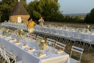 Outdoor wedding reception setup on a terrace with long white-draped tables, white chairs, and orange place card holders, overlooking a countryside landscape with a historic stone tower and trees.