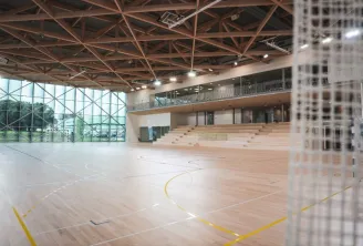 Modern sports hall interior at the University of Bordeaux with wooden flooring, marked court lines, exposed steel roof structure, large windows, and tiered seating area