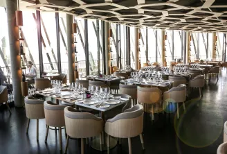 Modern panoramic restaurant interior with contemporary seating, tables set for dining, floor-to-ceiling windows overlooking Bordeaux cityscape, geometric ceiling design, and neutral-toned furnishings.
