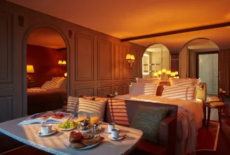 Luxury hotel suite at Mondrian Bordeaux Les Carmes featuring a breakfast table in the foreground, elegant beds with striped bedding, arched mirrors, warm ambient lighting, and modern furnishings in warm orange and burgundy tones.