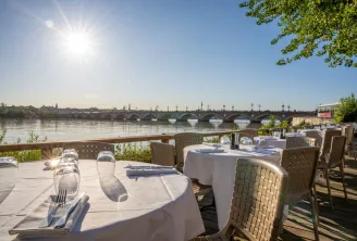Outdoor riverside restaurant terrace with white tablecloths overlooking a historic stone bridge spanning the Garonne River in Bordeaux on a sunny day