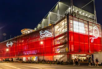 Night view of the UCPA Sport Station building in Bordeaux, featuring a modern red-lit facade with glass panels, metal framework, and illuminated upper levels with people visible on terraces.