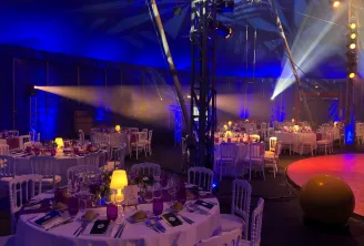 Elegant gala dinner setup in a large venue with round tables, white chairs, ambient blue and golden lighting, professional stage lighting rigs overhead, and a dance floor illuminated in red, ready for an evening event.