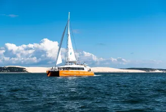Orange and white catamaran sailboat with white sails deployed, sailing in blue waters near white sand dunes and coastal cliffs under a clear sky with white clouds