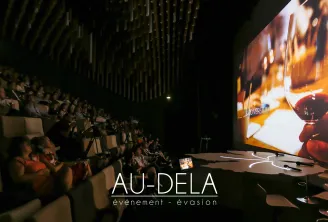 Indoor event venue with audience seated in tiered rows watching a large projection screen displaying amber-colored lighting and biodiversity content, with Au-Dela event company branding visible