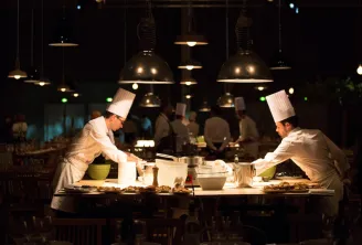 Two chefs in white uniforms and chef hats prepare food at a counter in a modern restaurant kitchen with hanging industrial lights, while diners are visible in the background.
