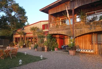 Modern industrial-style building with corrugated metal exterior in warm orange and red tones, featuring wooden balconies and terraces with potted plants, gravel courtyard, and green lawn area at dusk.