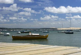 Golden rowing boat moored in turquoise waters of Arcachon Bay, with sailboats and sandy beach in foreground under blue sky with white clouds