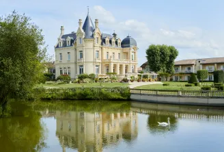 Grand Barrail castle hotel in Bordeaux, a cream-colored Victorian mansion with turrets and mansard roof reflected in a tranquil canal, surrounded by manicured gardens and greenery with a white swan on the water.