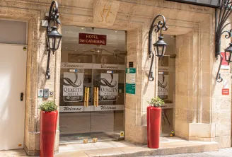 Entrance of Hotel Sainte Catherine, a Quality Hotel in Bordeaux city center, featuring glass doors with 'Welcome' and 'Bienvenue' text, decorative wrought iron lanterns, and red planters with greenery on the stone facade.
