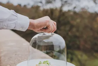 A chef's hand lifting a glass dome to reveal a gourmet plated dish with vegetables and garnish on a white plate, overlooking a snowy mountain landscape