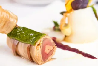 Elegant gourmet appetizer skewers featuring foie gras wrapped in bacon with green leaf garnish, served on a white plate with decorative elements