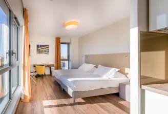 Modern hotel bedroom at All Suites Appart Hôtel Bordeaux Marne featuring a double bed with beige headboard, wooden flooring, large windows with city views, yellow accent curtains, desk area, and contemporary furnishings