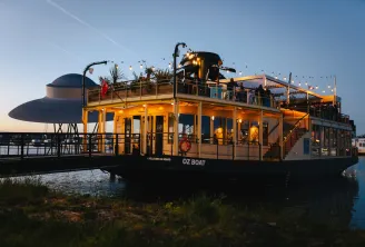 OZ Boat, an illuminated yellow river cruise boat docked at Bordeaux with a modern design, featuring multiple decks, outdoor terraces, and warm evening lighting against a twilight sky.