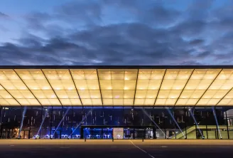 Modern illuminated building with a large translucent ceiling at dusk, labeled 'PALAIS 2' in Bordeaux, featuring contemporary architecture with glass facades and blue interior lighting.