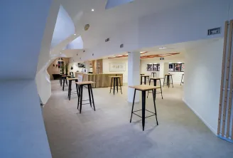 Modern interior of Club 33 at Mériadeck ice rink in Bordeaux, featuring high ceilings with skylights, contemporary bar seating with black stools and wooden tops, white walls, and event posters on display.