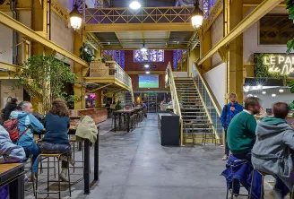Interior of a converted industrial space with yellow metal framework, featuring people seated at tables, brick walls, metal staircases, and ambient lighting in the evening.