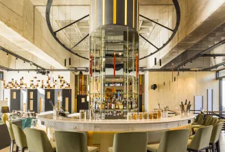 Modern hotel bar with a circular white counter, olive green bar stools, a striking black circular architectural element above, yellow and black striped central column, and geometric wall panels in a bright, contemporary space at FirstName JDV by Hyatt Bor