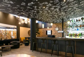 Modern hotel bar and lounge area with dark walls, textured ceiling, brick accent wall, pendant lighting, bar counter with stools, and comfortable seating with yellow and black accents