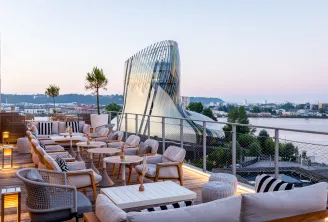 Rooftop terrace of Renaissance Bordeaux Hotel at dusk, featuring modern furniture, potted plants, and a panoramic view of the Garonne River and city skyline with an architectural sail-shaped building in the background.