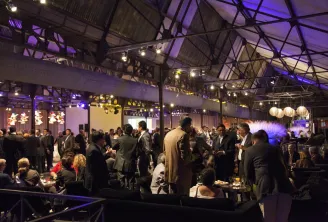 An elegant evening event at La Faïencerie in Bordeaux, featuring guests in formal attire mingling in a spacious industrial venue with purple ambient lighting, hanging lanterns, and exposed ceiling architecture.