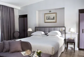 Modern luxury hotel bedroom at Hôtel de Sèze in Bordeaux, featuring a queen bed with gray upholstered headboard, white linens, twin nightstands with brass lamps, gray curtains, purple accent seating, and classical architectural framed print on the wall.