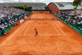 Professional tennis court with orange clay surface surrounded by spectators in stands, featuring BNP Paribas branding and traditional farm buildings in the background.