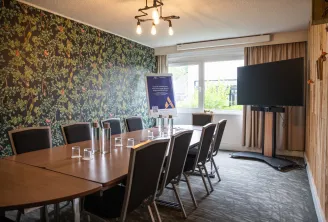 Modern conference room at Novotel Bordeaux Mérignac with wooden table, black chairs, floral patterned wallpaper, large display screen, and natural light from windows