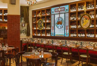 Elegant wine bar interior with wooden shelving displaying hundreds of wine bottles, red velvet banquette seating, round tables, ornate decorative panels, and vintage wine-themed artwork on the walls.