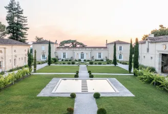 Elegant neoclassical estate with white stone buildings surrounding a manicured courtyard featuring a central rectangular pool, cypress trees, and manicured lawns at dusk in Bordeaux.