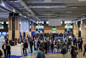 Large indoor convention center or trade show in Bordeaux with numerous attendees in business attire circulating among exhibition booths and information stands under modern ceiling lighting.