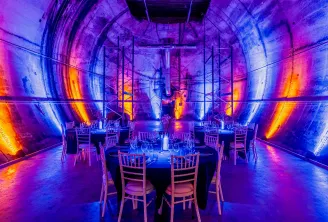 A modern dining space inside an industrial tunnel illuminated with vibrant neon lighting in shades of blue, purple, and orange. Round tables set with place settings and chairs are arranged symmetrically along the tunnel, with metal framework visible overh