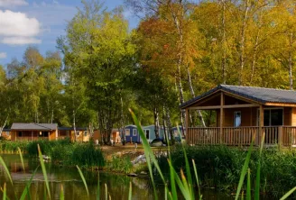 Lakeside camping village with wooden chalets and colorful holiday homes surrounded by birch trees and reeds, reflected in calm water under a partly cloudy sky