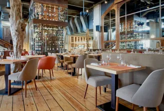 Modern restaurant interior with wooden floors, comfortable beige and red chairs, tables with place settings, extensive bottle display behind glass, and industrial ceiling with exposed beams in Bordeaux Maritime Café.