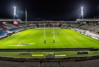 Stade Matmut Bordeaux rugby field at night, illuminated with floodlights, showing the white lines of the rugby pitch with two goal posts centered, surrounded by tiered seating in the stadium.