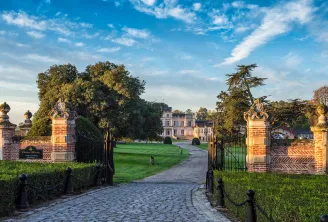 Grand entrance gates of Château Giscours in Bordeaux, with ornate stone pillars flanking a tree-lined driveway leading to a pink neoclassical mansion under a blue sky.