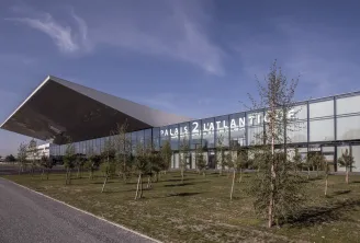 Modern glass and steel exhibition hall with 'Palais 2 L'Atlantique' signage, surrounded by young trees and landscaping in Bordeaux.