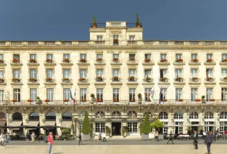 The Intercontinental Grand Hôtel de Bordeaux, an elegant 18th-century neoclassical building with cream-colored stone facade, ornate balconies, and mansard roofs with golden spires, facing a public plaza in downtown Bordeaux.