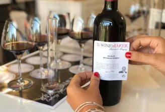 A person holding a bottle of B.Winemaker wine by Bernard Magrez, showing a blend of 15% Merlot and 85% Cabernet Sauvignon, with wine glasses and place settings visible on a dining table in the background.