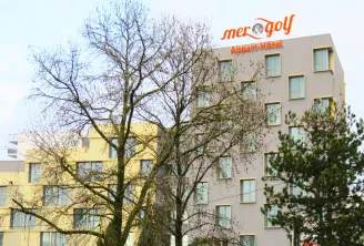 Exterior view of Mer & Golf Aparthotel, a modern multi-story building with beige and yellow facades, featuring regular window arrangements and the hotel signage on top, surrounded by bare trees in winter.