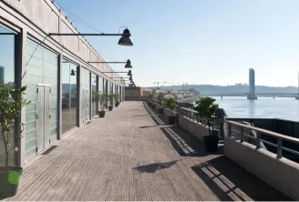 Modern waterfront terrace at Hangar 14 in Bordeaux, featuring a wooden deck with white buildings, black pendant lights, potted plants, and a view of the Garonne River with a bridge in the background under clear skies.
