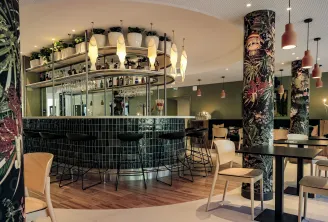 Modern hotel bar and lounge at Ibis Styles Bordeaux Lac Bruges featuring a curved black tiled bar counter with illuminated pendant lights, decorative botanical-patterned columns, comfortable seating areas, and potted plants throughout the space.
