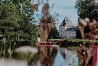 A picturesque view of Château Larrivet Haut-Brion in Bordeaux, featuring a pink arched bridge over a calm water canal, surrounded by lush gardens with blooming flowers, tall trees, and the château's distinctive mansard-roofed architecture visible in the b